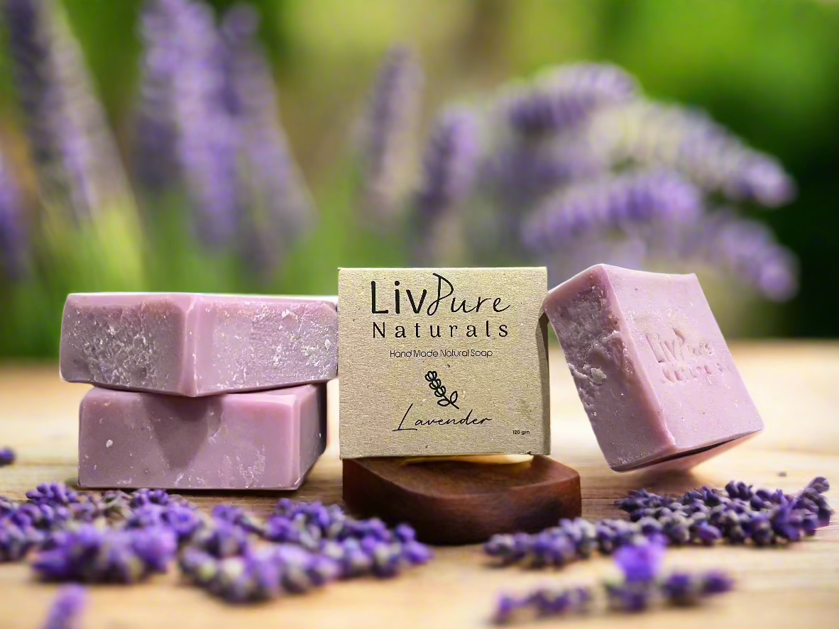 Handmade lavender-colored soap bars with a label that reads 'LivPure Naturals - Lavender', displayed on a wooden surface with lavender flowers scattered around and a blurred background.